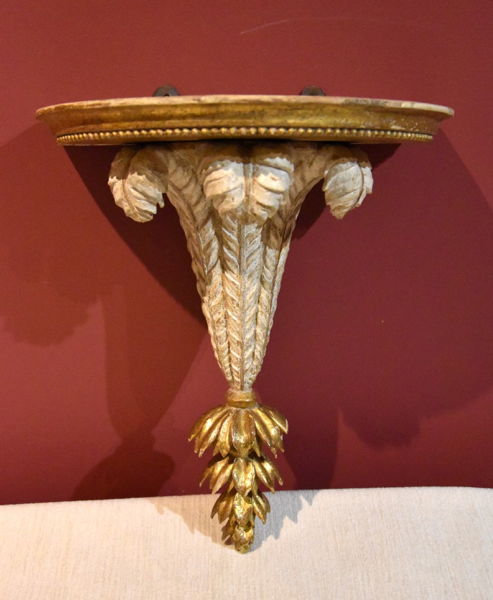 An 18th-century carved wall bracket featuring a semicircular gilded top shelf with beaded molding above a tapered white body with leaf carving and a drop leaf finial. View II