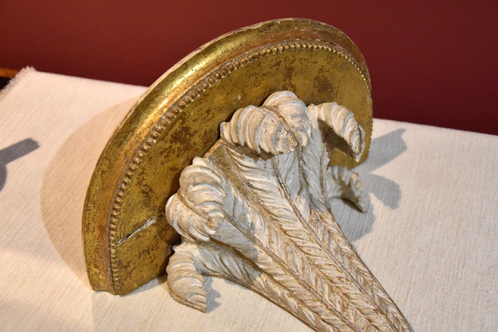 An 18th-century carved wall bracket featuring a semicircular gilded top shelf with beaded molding above a tapered white body with leaf carving and a drop leaf finial. View VI