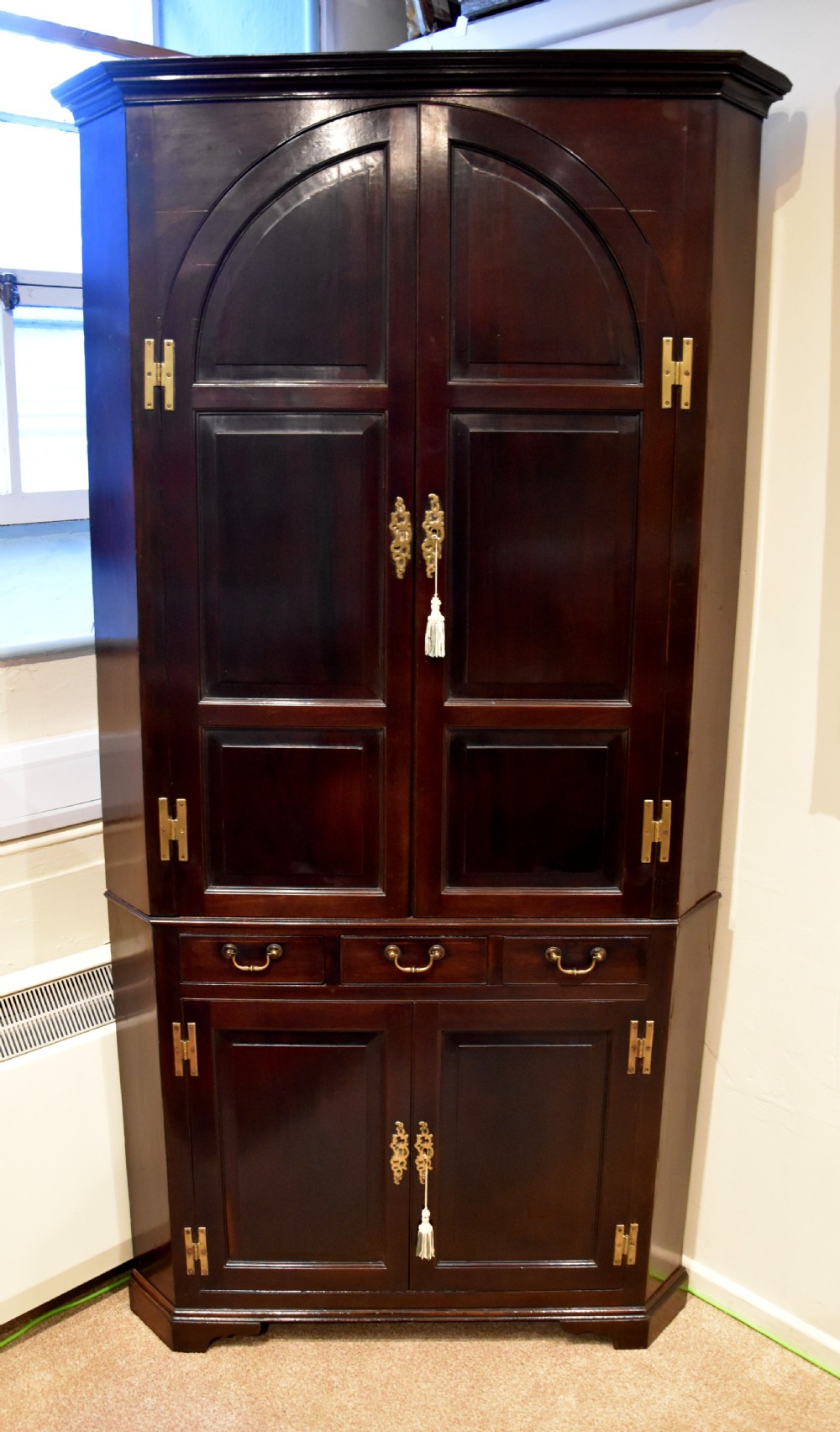An 18th-century Cuban mahogany tall corner cupboard featuring arched panel doors, brass H-hinges, middle drawers, and lower cabinet doors.