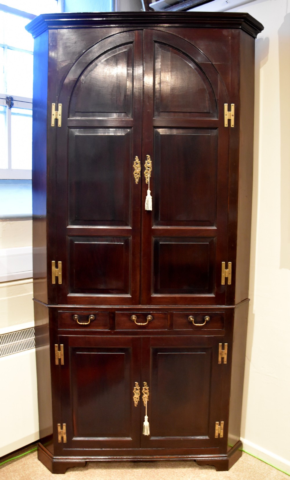 An 18th-century Cuban mahogany tall corner cupboard featuring arched panel doors, brass H-hinges, middle drawers, and lower cabinet doors. View II