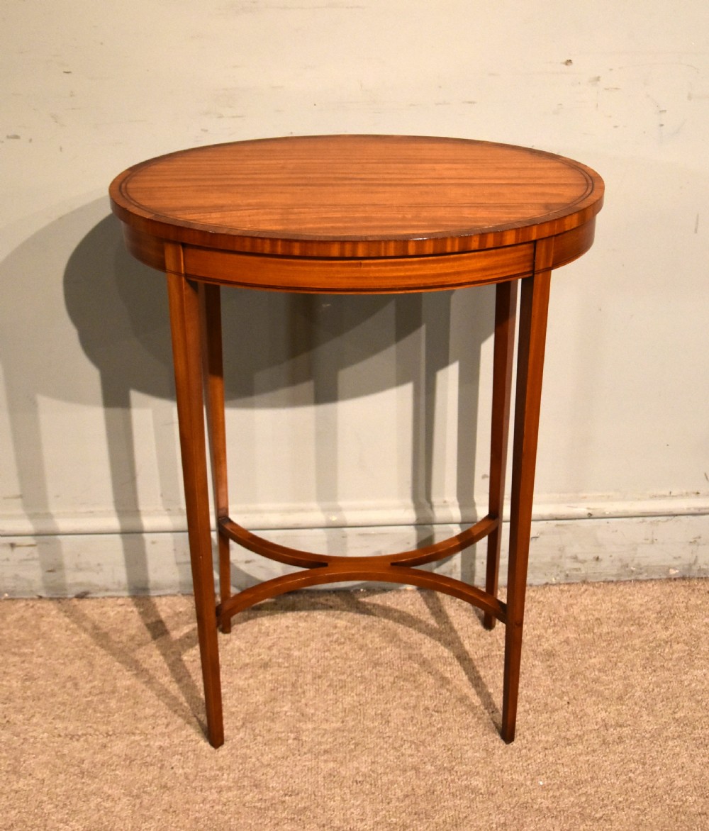 An oval Sheraton Revival occasional table crafted from satinwood with crossbanded border inlay, slender tapered legs, and a curved X-frame stretcher.