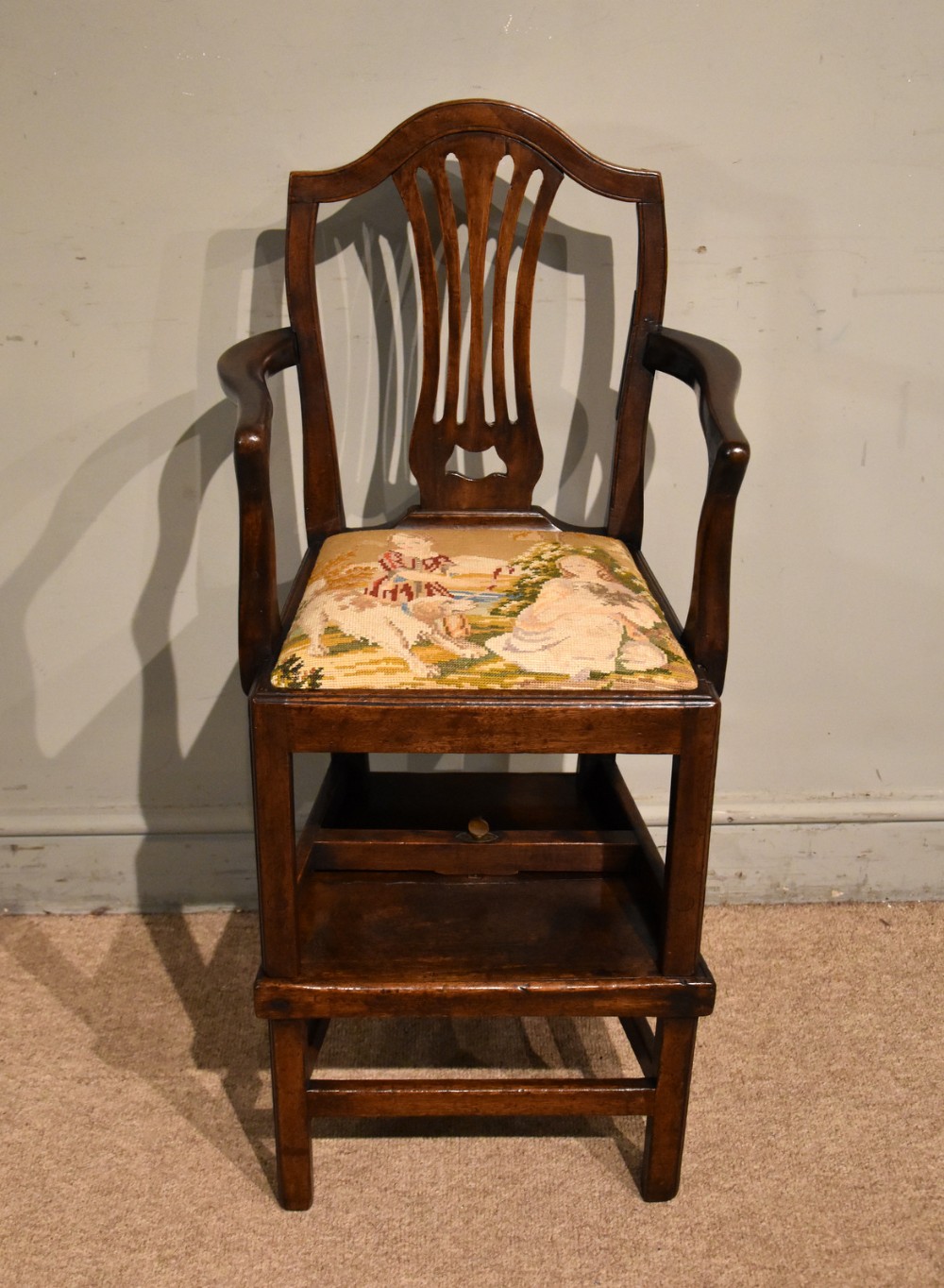 An antique dark wood George III convertible highchair featuring a hand-carved pierced back splat, curved armrests, and a tapestry upholstered seat depicting figures in a landscape.