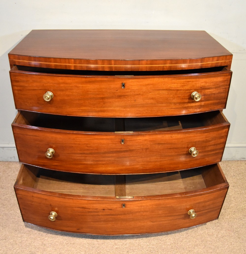A Regency period mahogany bow-fronted chest featuring three full-width drawers with brass handles and splayed bracket feet. View V