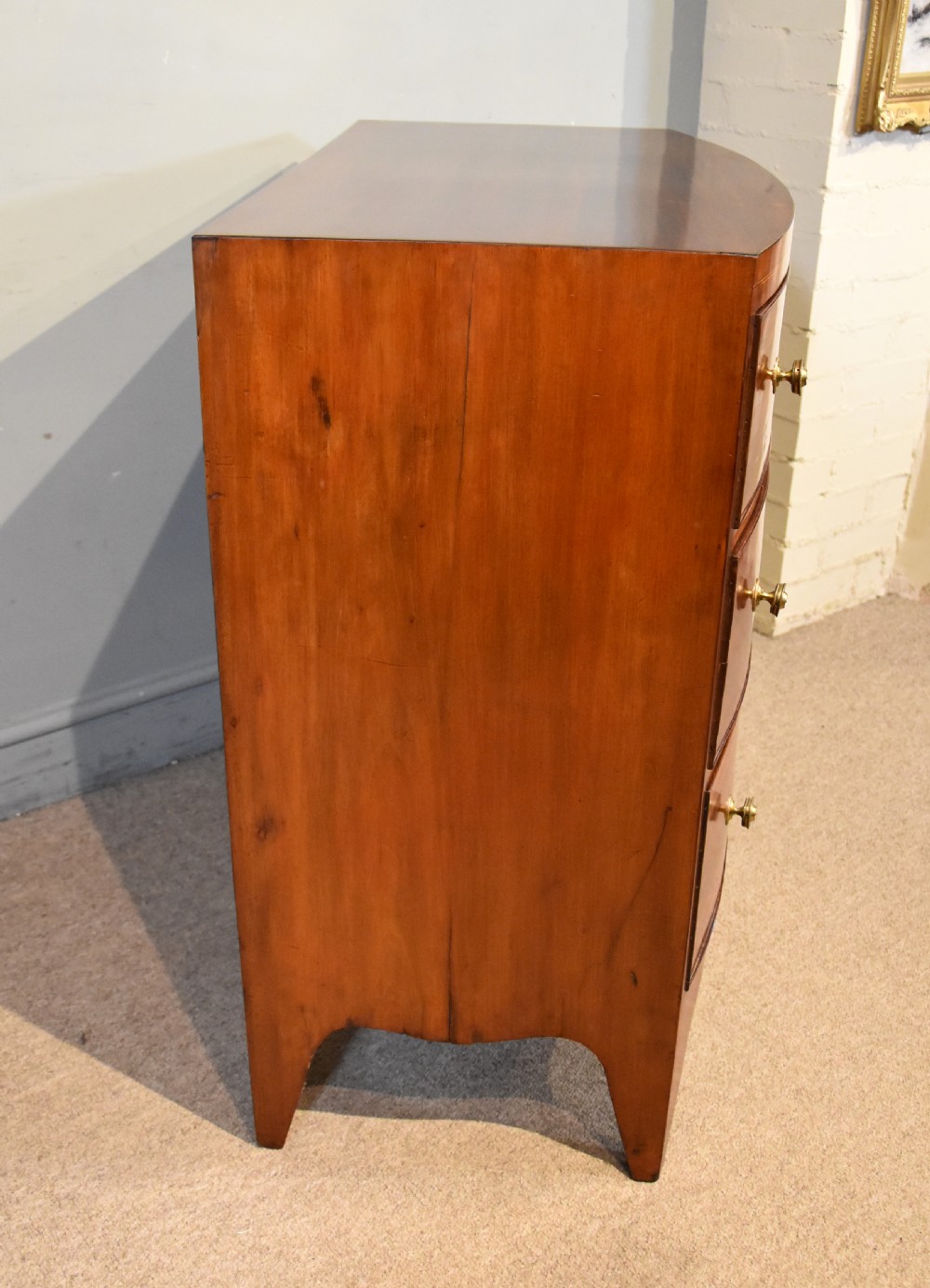 A Regency period mahogany bow-fronted chest featuring three full-width drawers with brass handles and splayed bracket feet. View VII