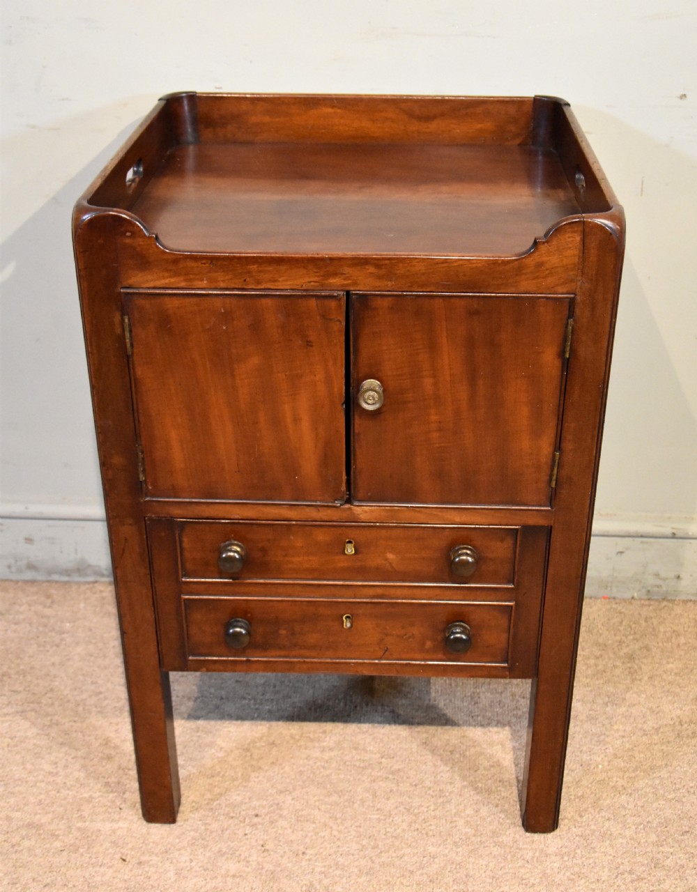 A George III mahogany bedside cabinet featuring a gallery tray top with pierced side handles, double cupboard doors, and two drawers below.
