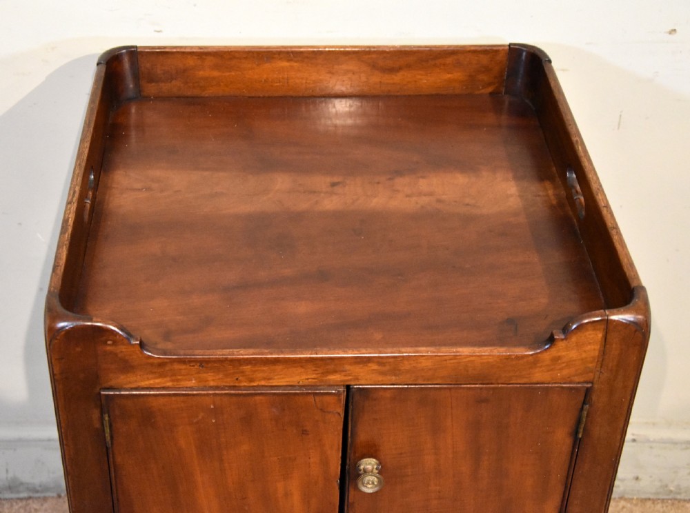 A George III mahogany bedside cabinet featuring a gallery tray top with pierced side handles, double cupboard doors, and two drawers below. View IV