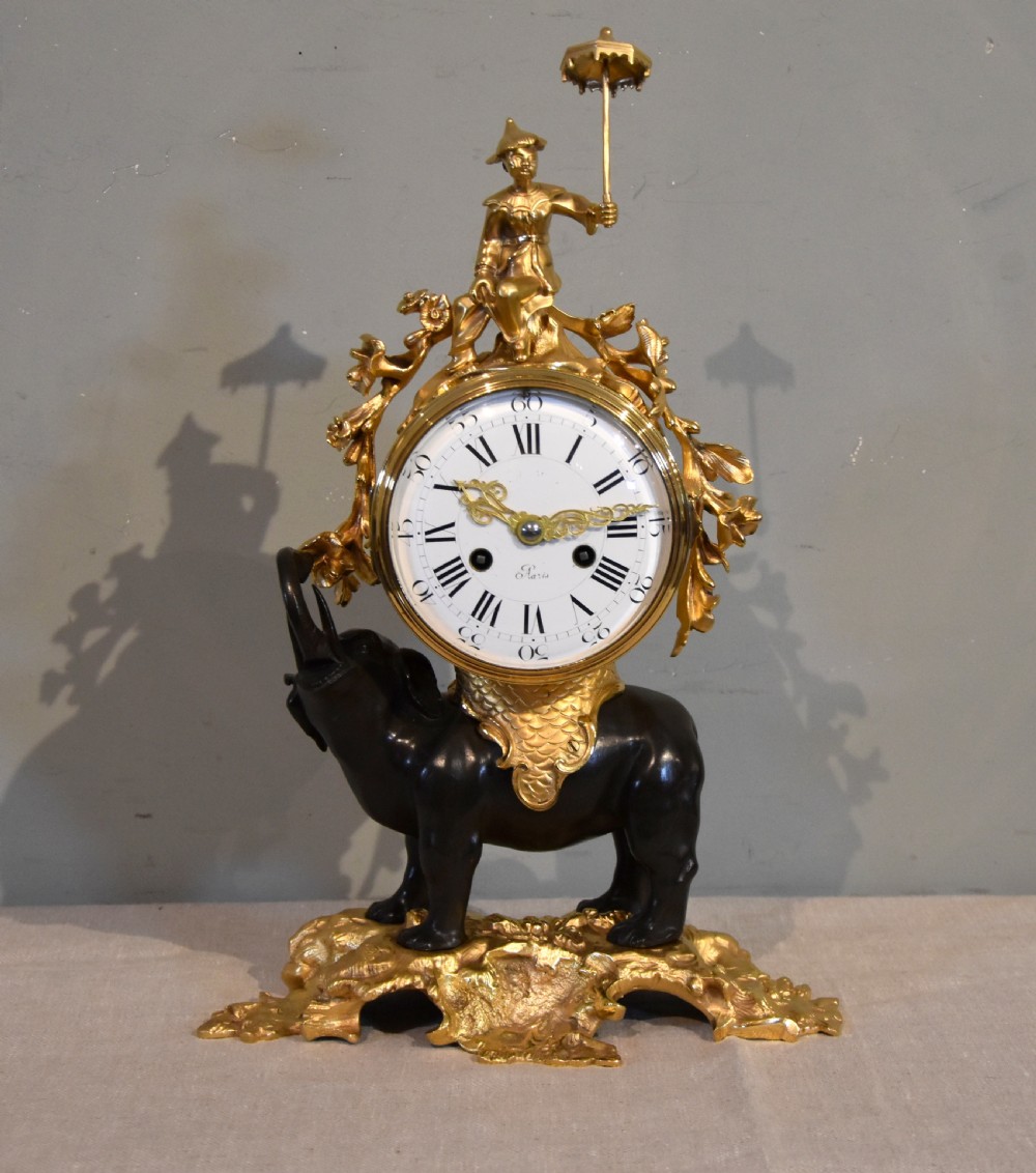 A French Louis XV style mantel clock featuring a dark patinated bronze elephant supporting an ormolu clock drum topped with a figure holding a parasol, mounted on a gilded rocaille base.