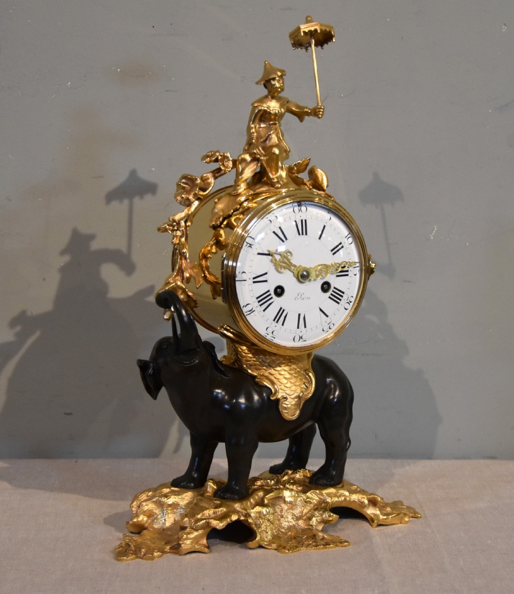 A French Louis XV style mantel clock featuring a dark patinated bronze elephant supporting an ormolu clock drum topped with a figure holding a parasol, mounted on a gilded rocaille base. View II
