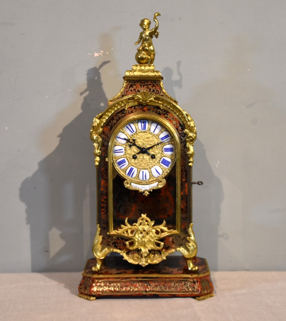 An ornate Boulle work mantel clock featuring red tortoiseshell and brass marquetry, topped with a gilt bronze cherub figure and set with blue enamel Roman numeral cartouches.