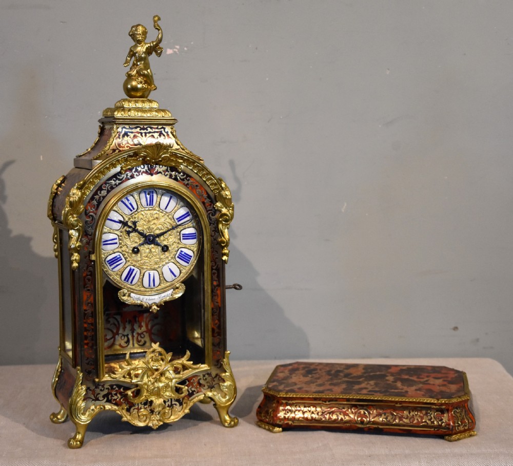An ornate Boulle work mantel clock featuring red tortoiseshell and brass marquetry, topped with a gilt bronze cherub figure and set with blue enamel Roman numeral cartouches. View II