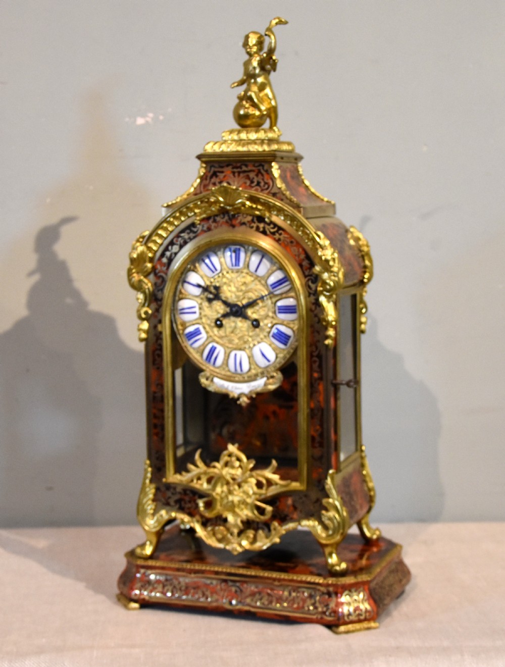 An ornate Boulle work mantel clock featuring red tortoiseshell and brass marquetry, topped with a gilt bronze cherub figure and set with blue enamel Roman numeral cartouches. View III