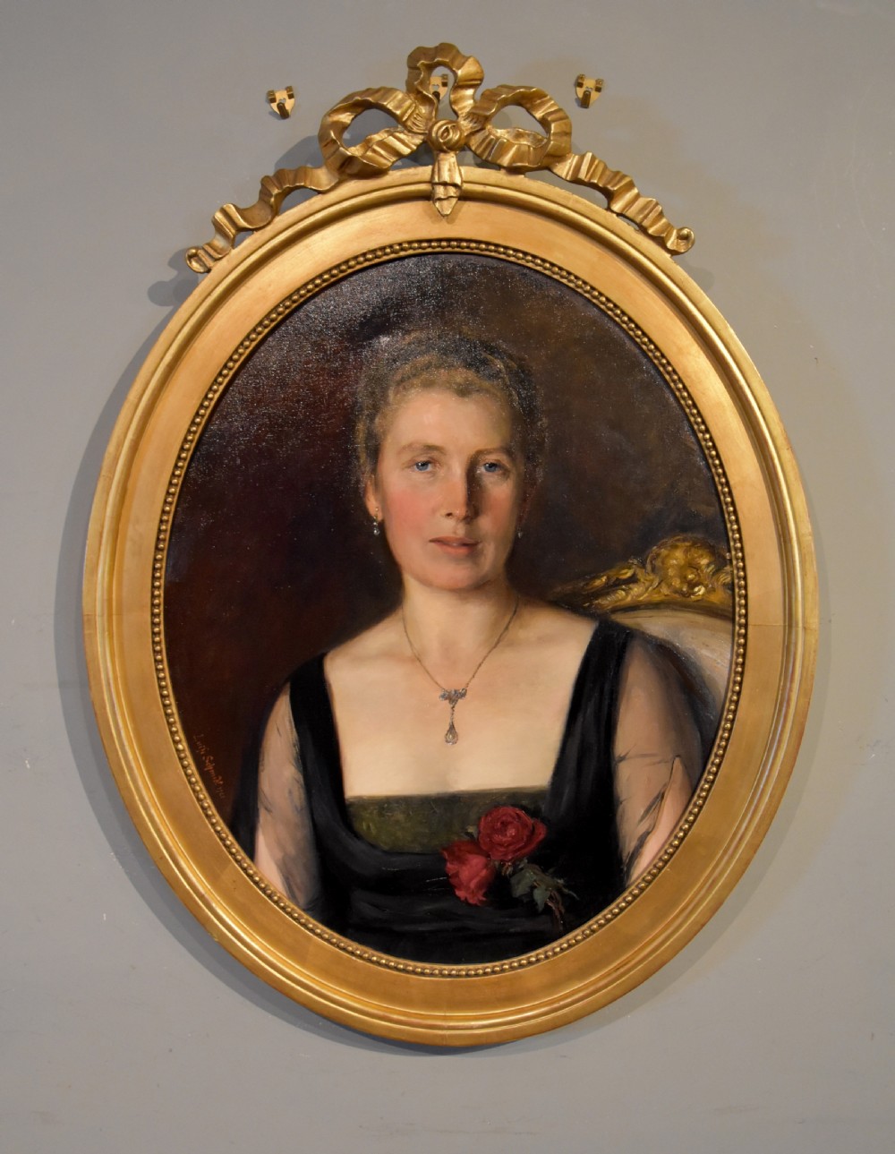 An oval antique oil painting featuring a seated lady in a black gown with a red rose corsage, set within a carved giltwood frame with a ribbon cresting.