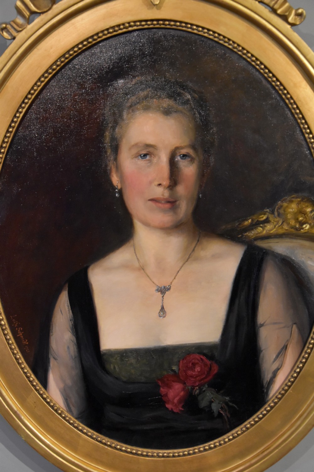 An oval antique oil painting featuring a seated lady in a black gown with a red rose corsage, set within a carved giltwood frame with a ribbon cresting. View II