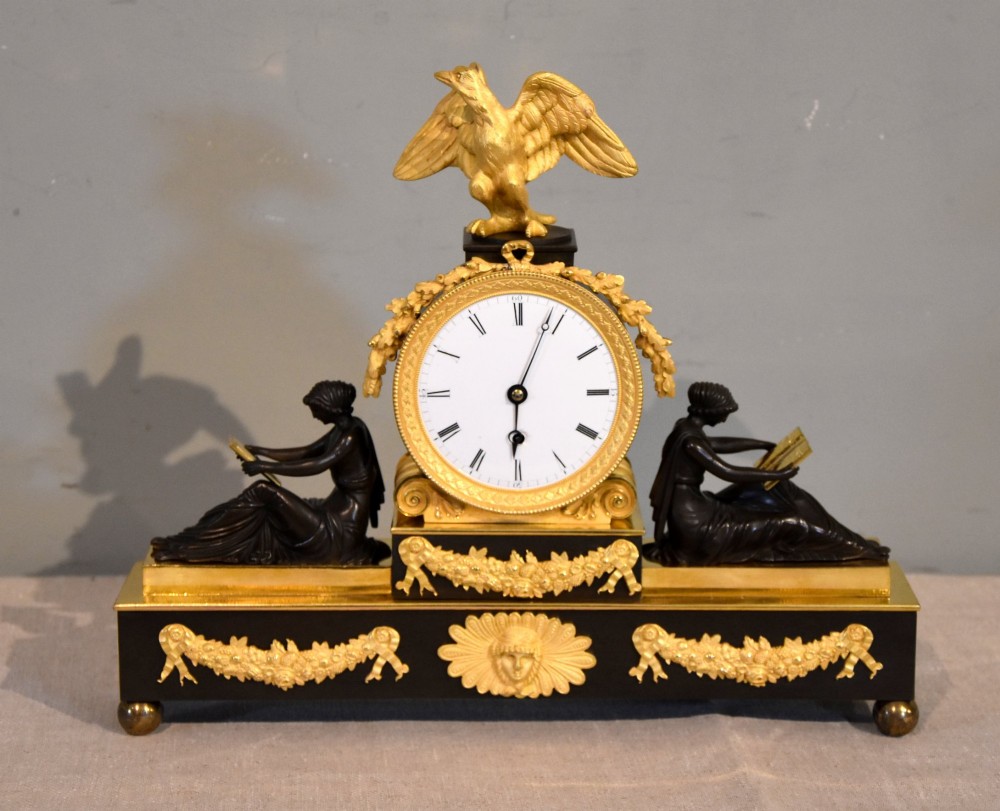 An English Regency gilt brass and patinated bronze mantel timepiece featuring a top eagle finial, circular enamel dial, and two seated classical female figures.
