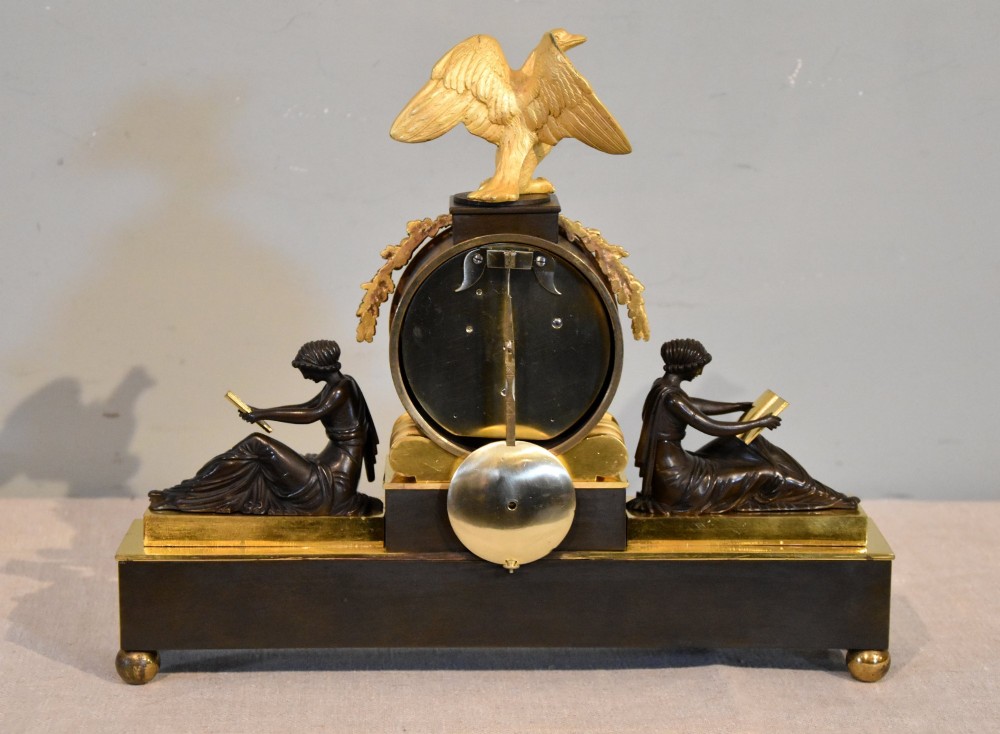 An English Regency gilt brass and patinated bronze mantel timepiece featuring a top eagle finial, circular enamel dial, and two seated classical female figures. View V