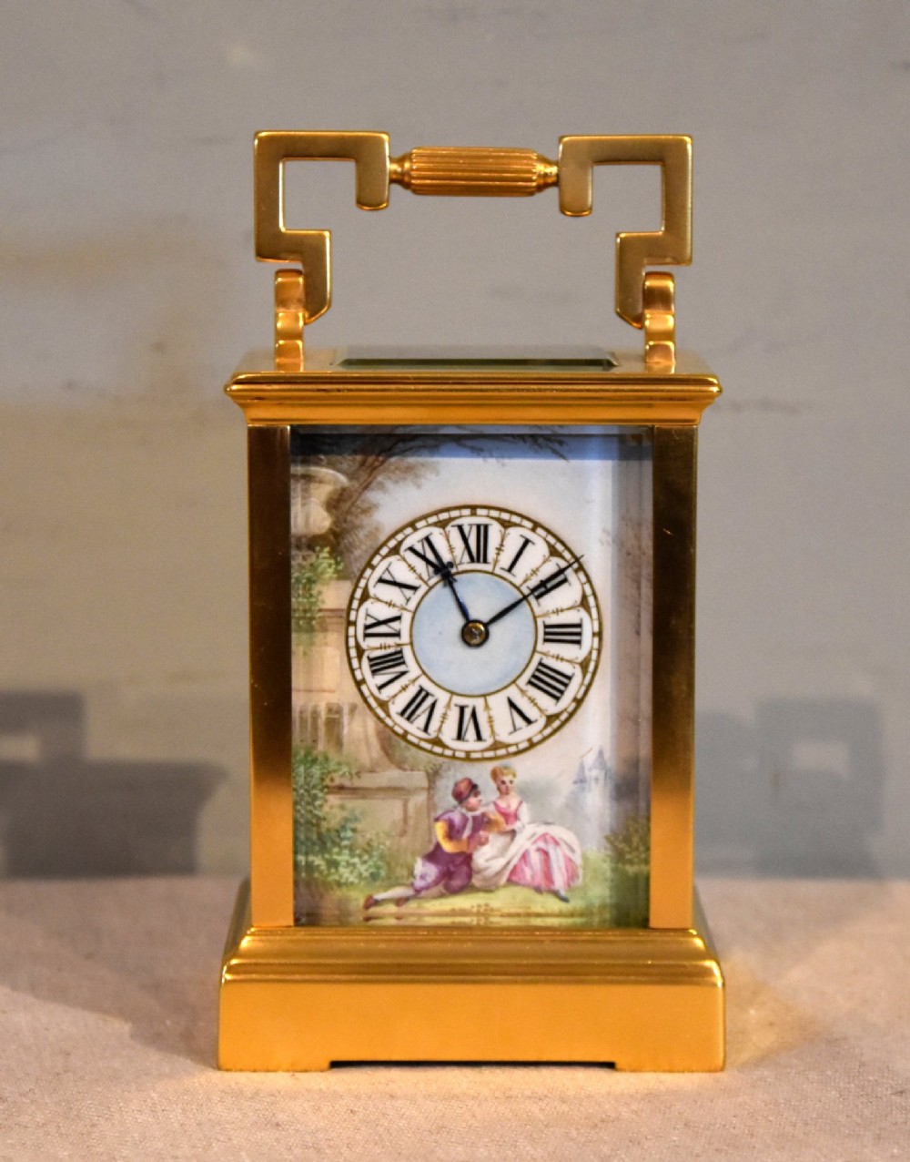 A gilt brass carriage clock featuring a rectangular case, a geometric top handle, and a hand-painted porcelain front panel illustrating a romantic couple in a landscape.