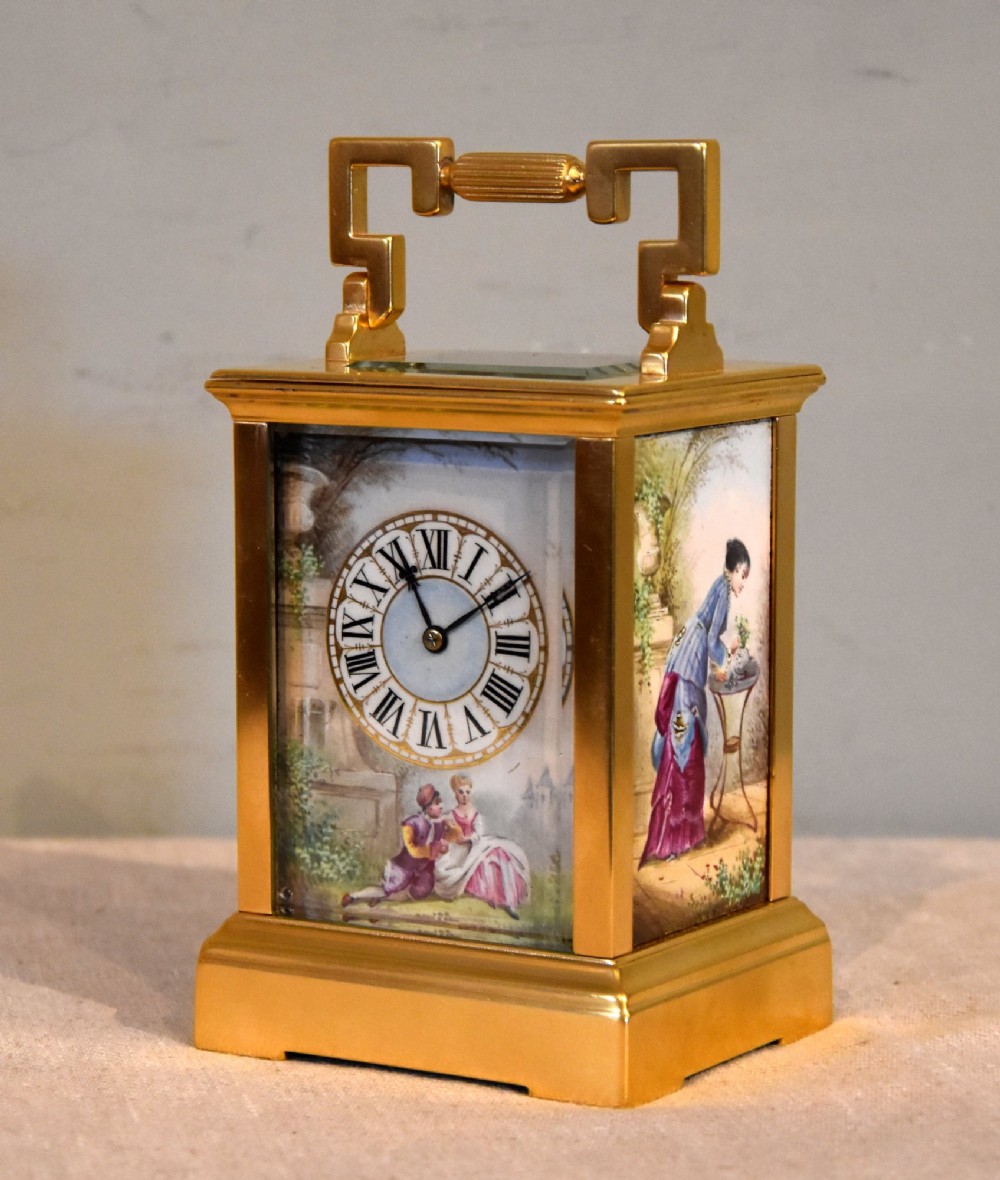 A gilt brass carriage clock featuring a rectangular case, a geometric top handle, and a hand-painted porcelain front panel illustrating a romantic couple in a landscape. View II