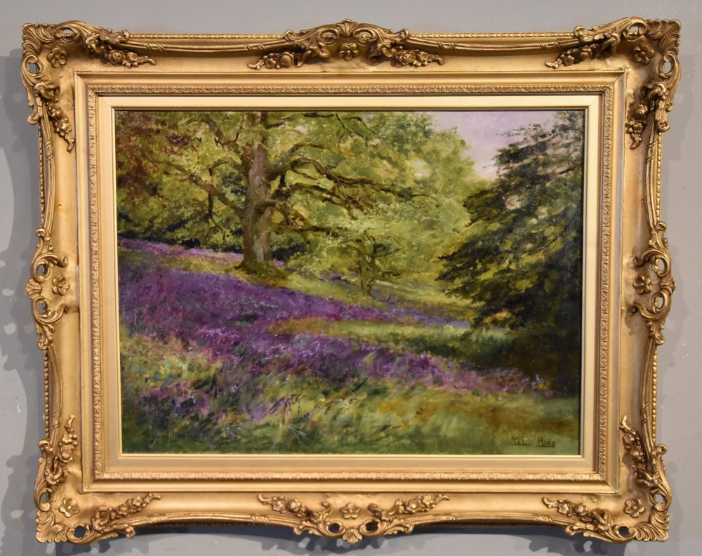 An antique oil painting depicting a woodland landscape with purple flowers beneath a tree, displayed within an ornate gilt frame.