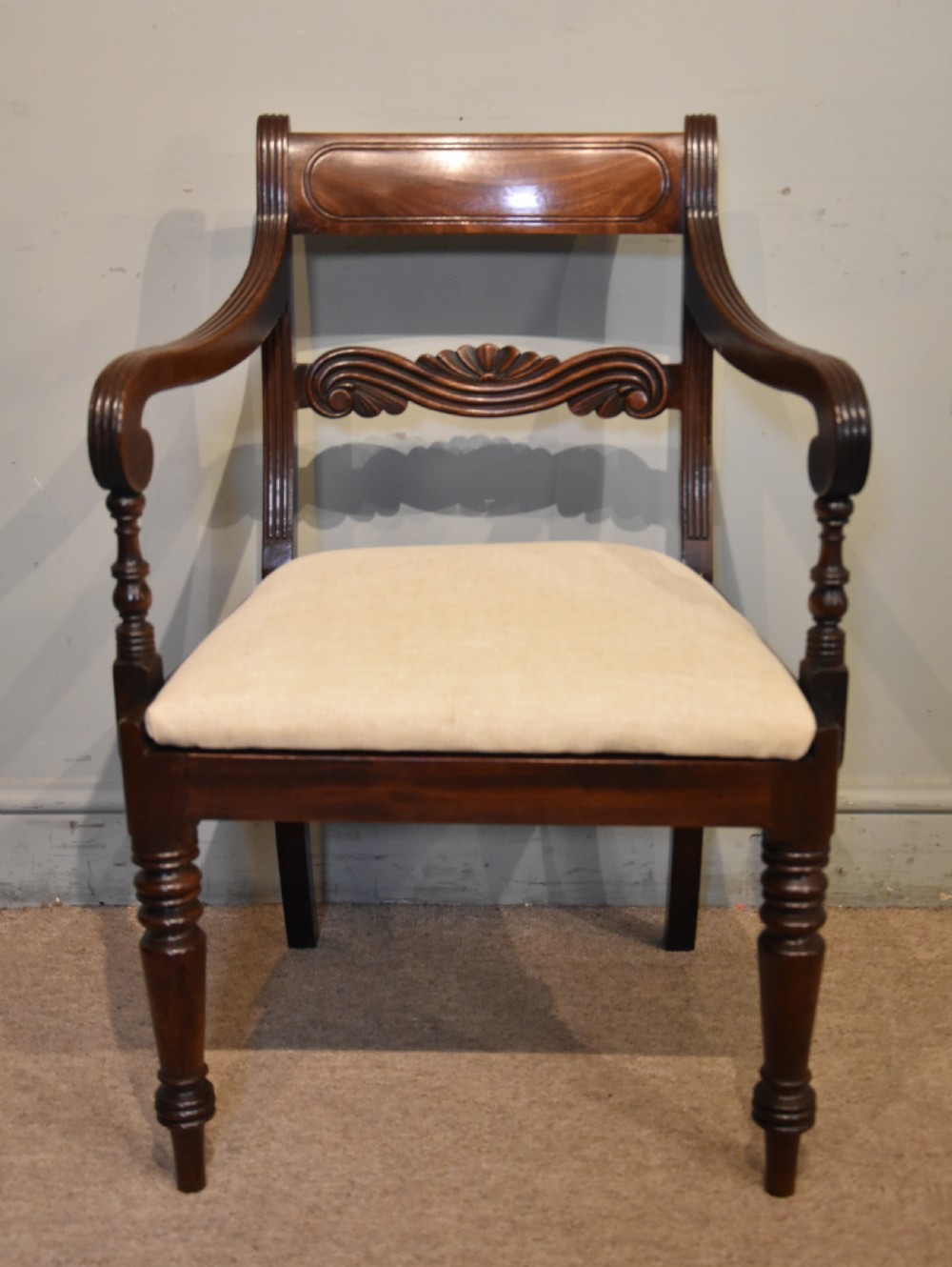 A Regency period mahogany open armchair featuring a reeded tablet top rail, carved scroll mid-rail, turned front legs, and cream fabric seat.