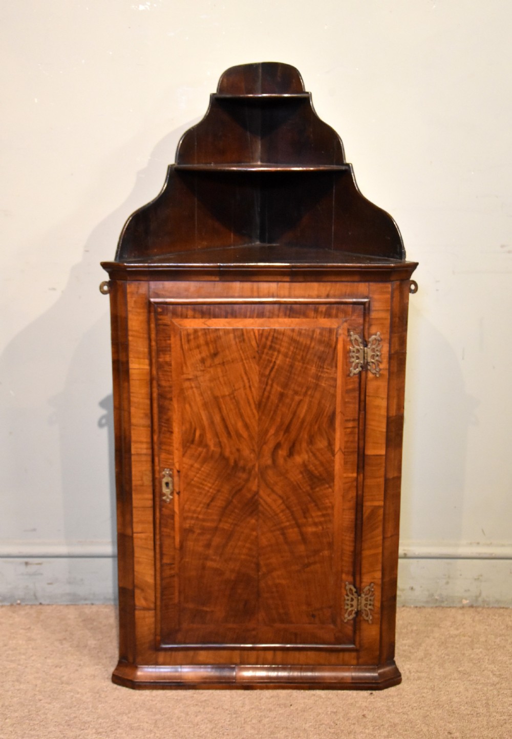 A George I walnut corner cupboard featuring a tiered open shelf superstructure above a panelled door with book-matched veneer and brass hinges.