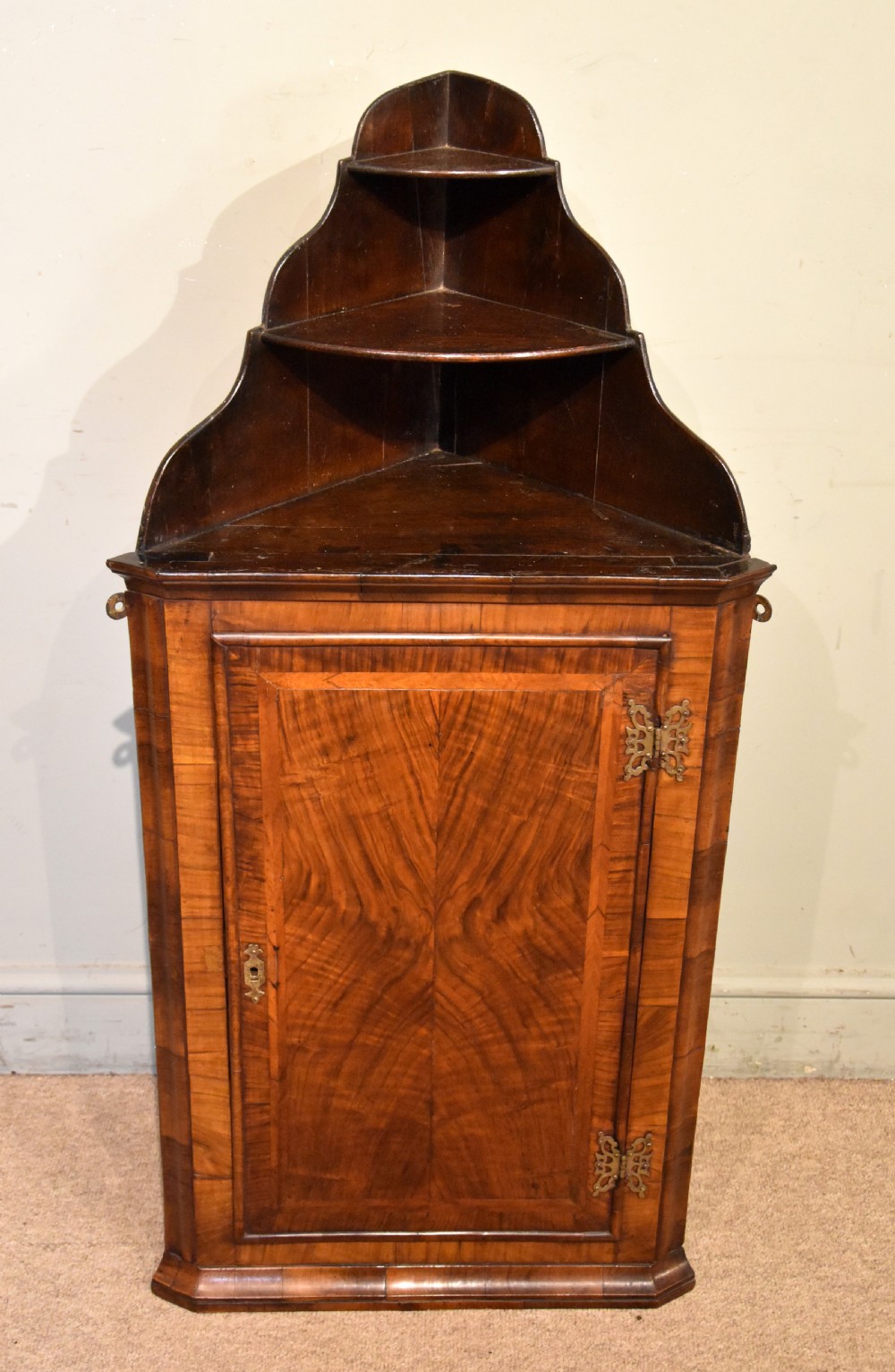 A George I walnut corner cupboard featuring a tiered open shelf superstructure above a panelled door with book-matched veneer and brass hinges. View II