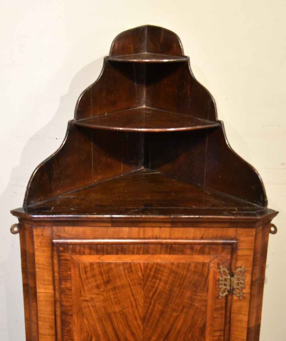 A George I walnut corner cupboard featuring a tiered open shelf superstructure above a panelled door with book-matched veneer and brass hinges. View V