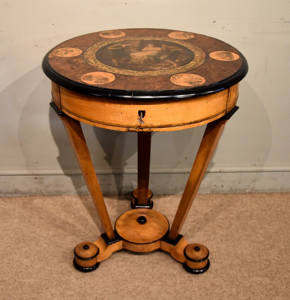 A circular Biedermeier centre table featuring a painted classical top panel, golden birch veneer apron with key, ebonised trim, and a tripod base.