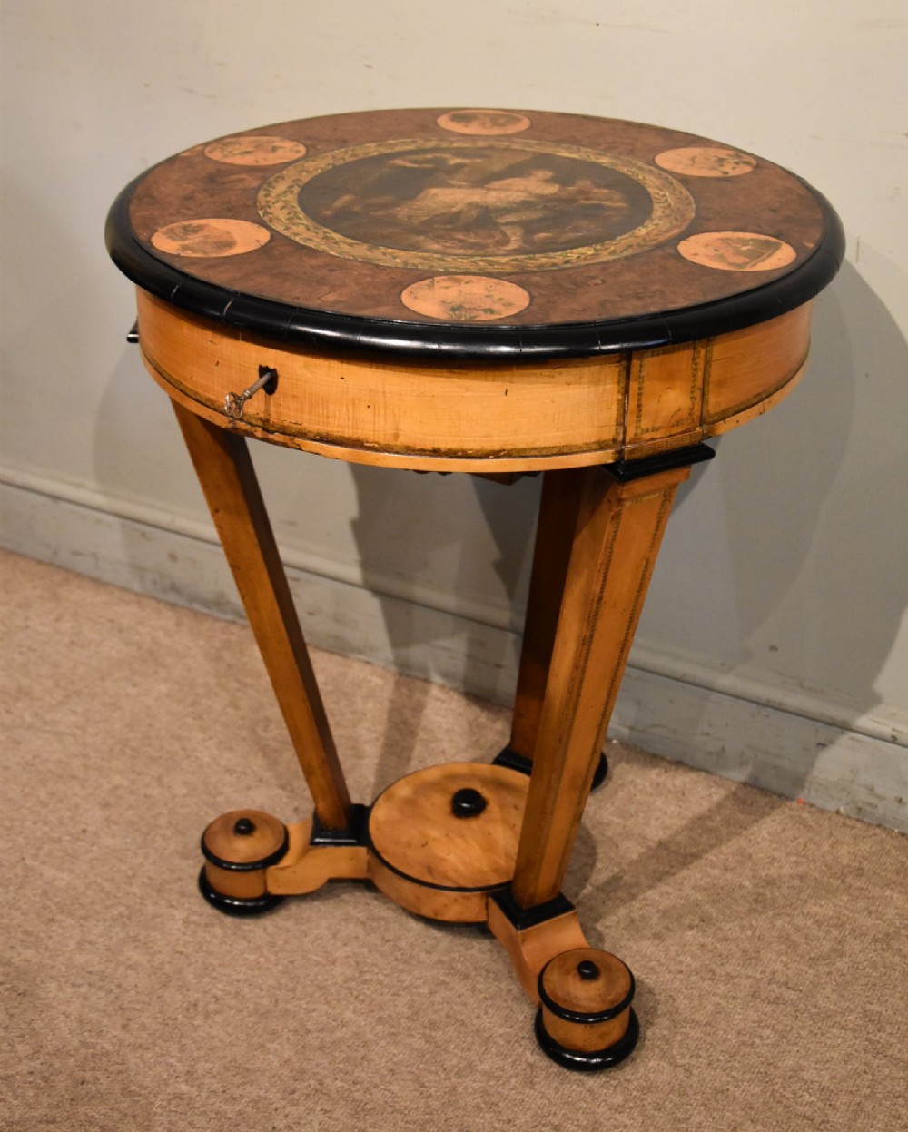 A circular Biedermeier centre table featuring a painted classical top panel, golden birch veneer apron with key, ebonised trim, and a tripod base. View VII