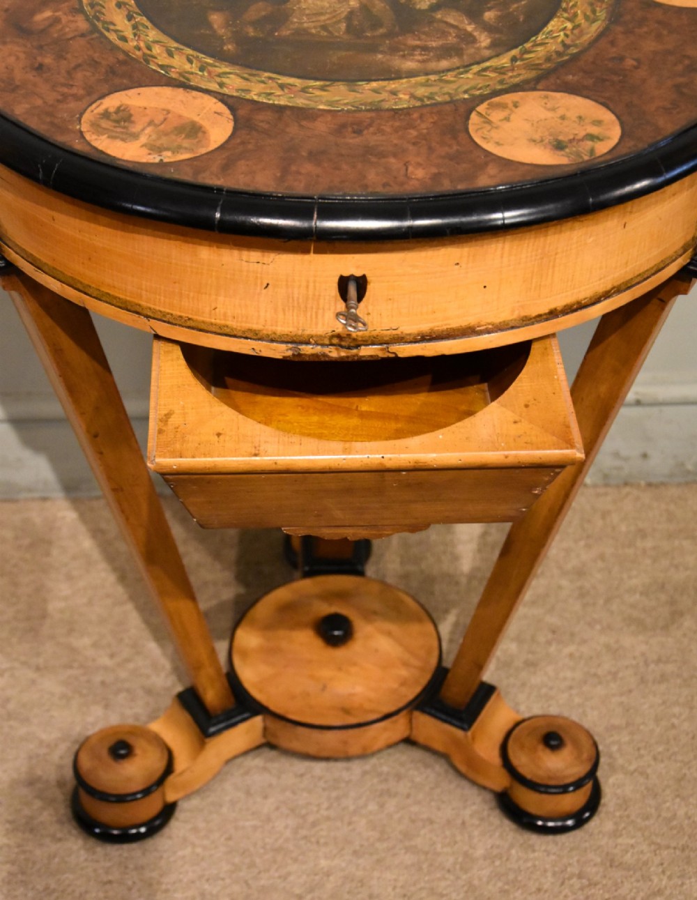 A circular Biedermeier centre table featuring a painted classical top panel, golden birch veneer apron with key, ebonised trim, and a tripod base. View IX