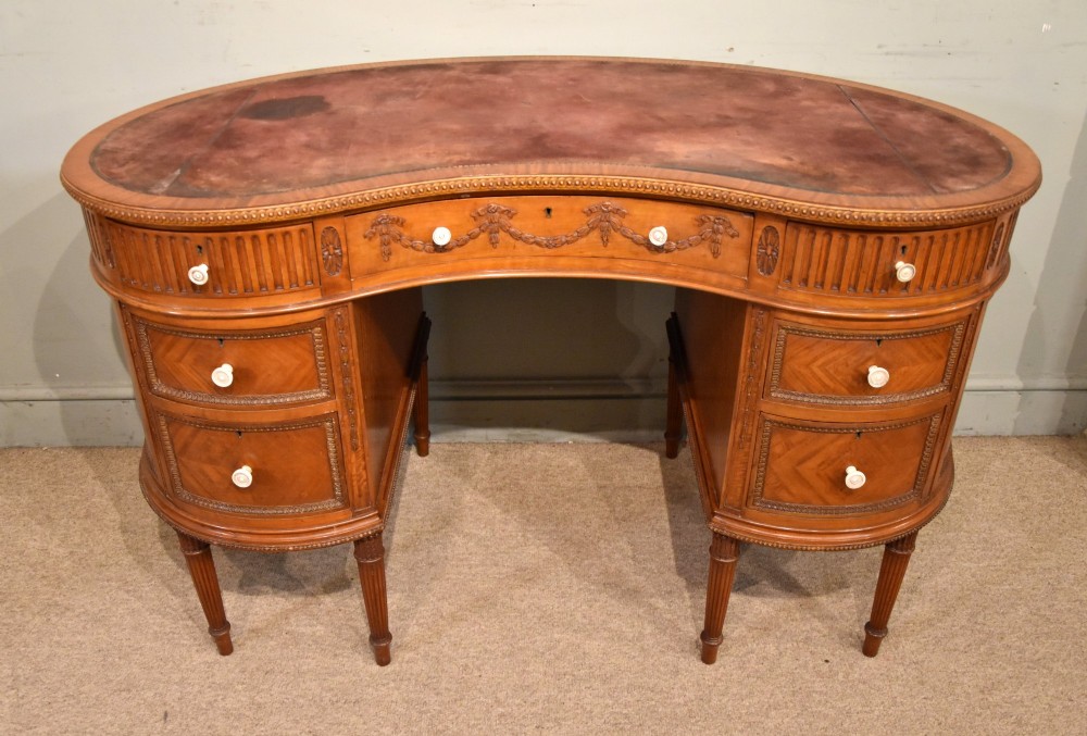 An Edwardian satinwood kidney-shaped writing desk featuring an inset leather top, carved floral swags, and five drawer fronts on fluted legs.