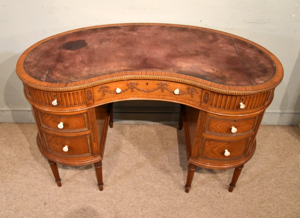An Edwardian satinwood kidney-shaped writing desk featuring an inset leather top, carved floral swags, and five drawer fronts on fluted legs. View II