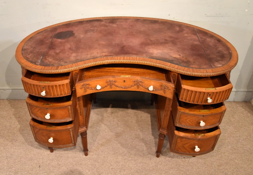 An Edwardian satinwood kidney-shaped writing desk featuring an inset leather top, carved floral swags, and five drawer fronts on fluted legs. View III