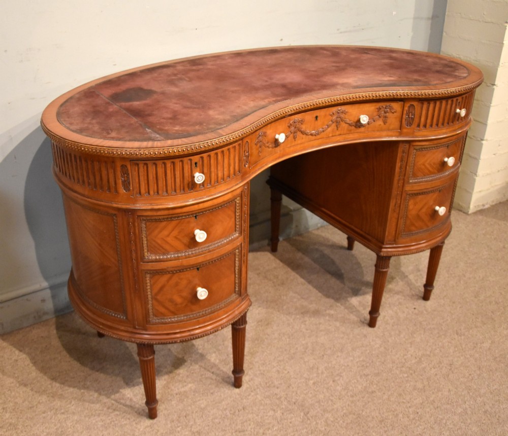 An Edwardian satinwood kidney-shaped writing desk featuring an inset leather top, carved floral swags, and five drawer fronts on fluted legs. View IV