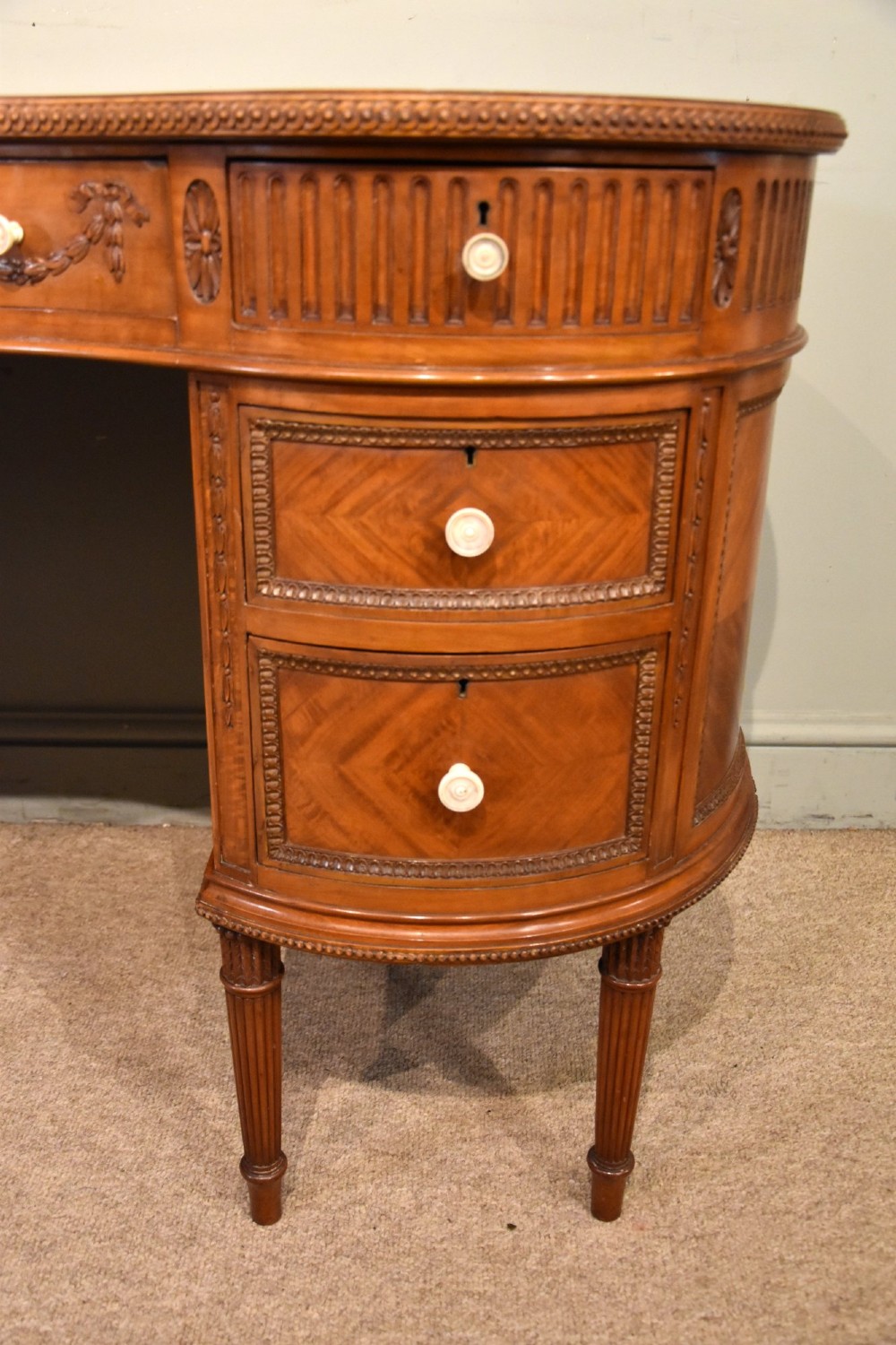 An Edwardian satinwood kidney-shaped writing desk featuring an inset leather top, carved floral swags, and five drawer fronts on fluted legs. View V