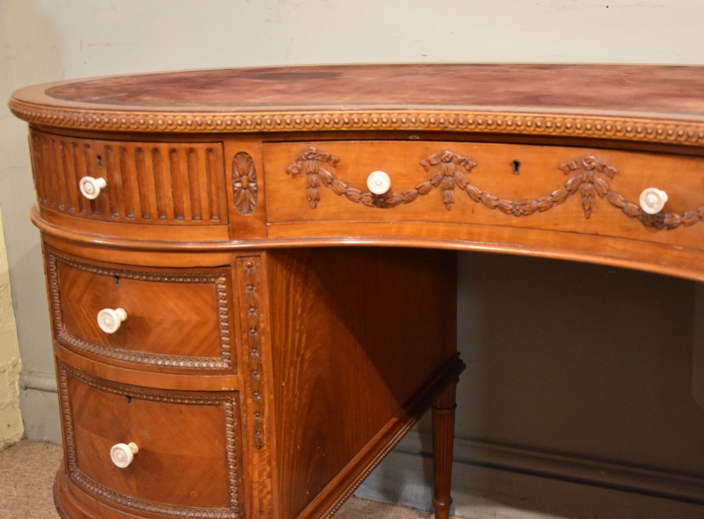 An Edwardian satinwood kidney-shaped writing desk featuring an inset leather top, carved floral swags, and five drawer fronts on fluted legs. View VI
