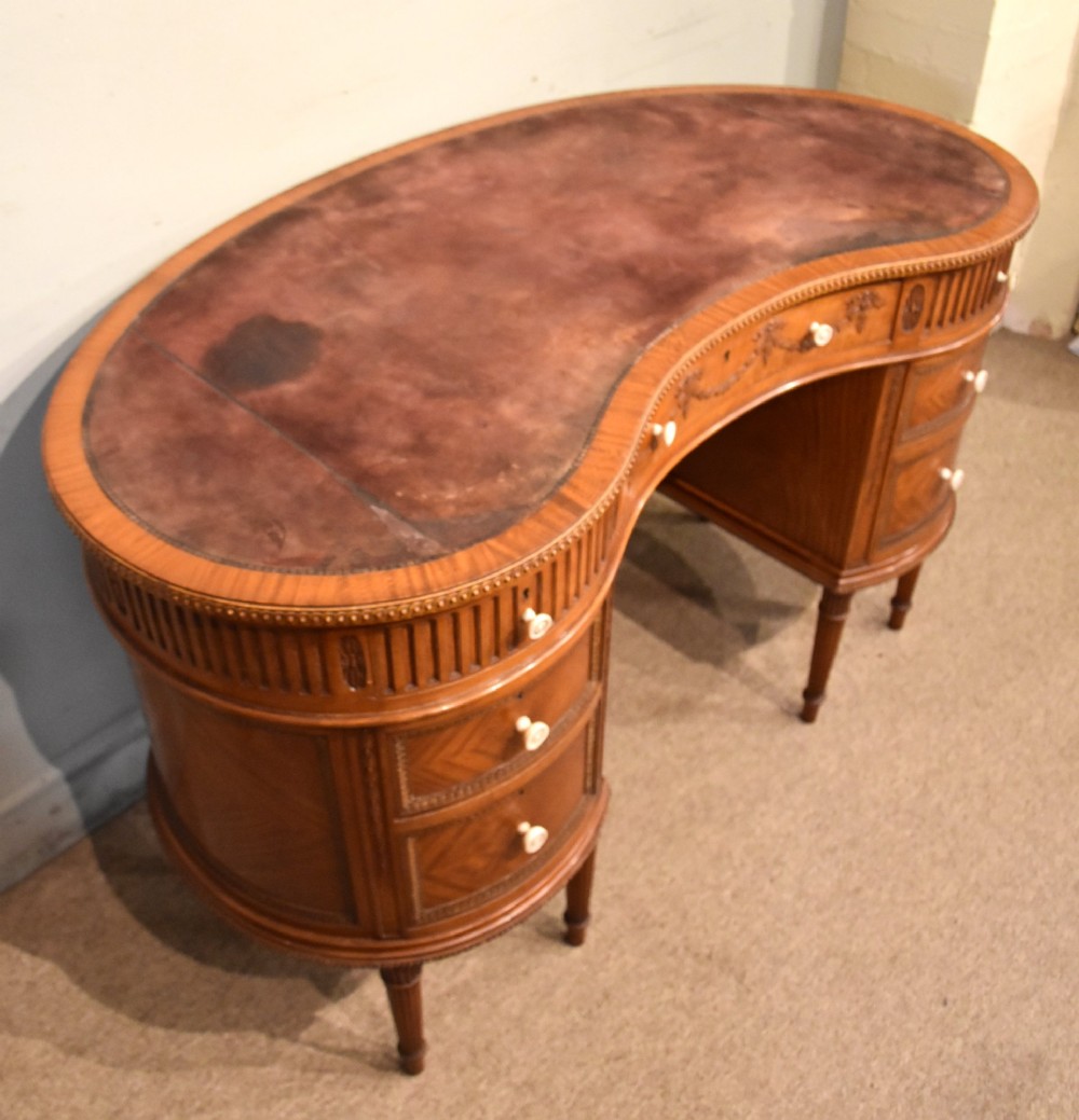 An Edwardian satinwood kidney-shaped writing desk featuring an inset leather top, carved floral swags, and five drawer fronts on fluted legs. View VII