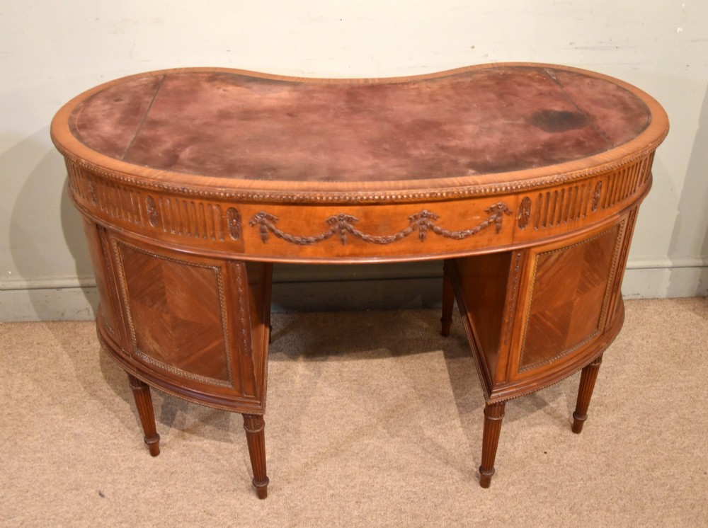 An Edwardian satinwood kidney-shaped writing desk featuring an inset leather top, carved floral swags, and five drawer fronts on fluted legs. View VIII