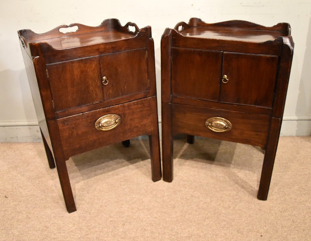 A matched pair of dark mahogany Georgian bedside cabinets, each featuring a shaped gallery top with cutout handles, a two-door cabinet, and a single lower drawer with a brass handle.