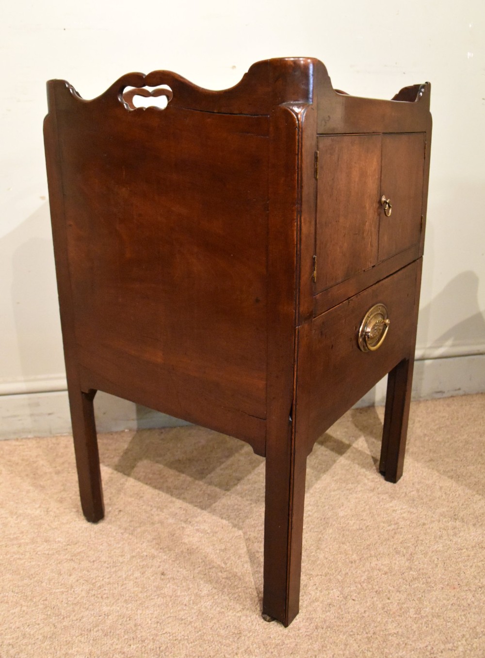 A matched pair of dark mahogany Georgian bedside cabinets, each featuring a shaped gallery top with cutout handles, a two-door cabinet, and a single lower drawer with a brass handle. View VII