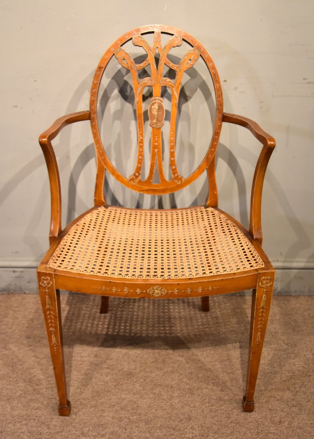 An Edwardian satinwood open armchair featuring an oval pierced backrest, hand-painted floral motifs, sweeping arms, and a woven cane seat.