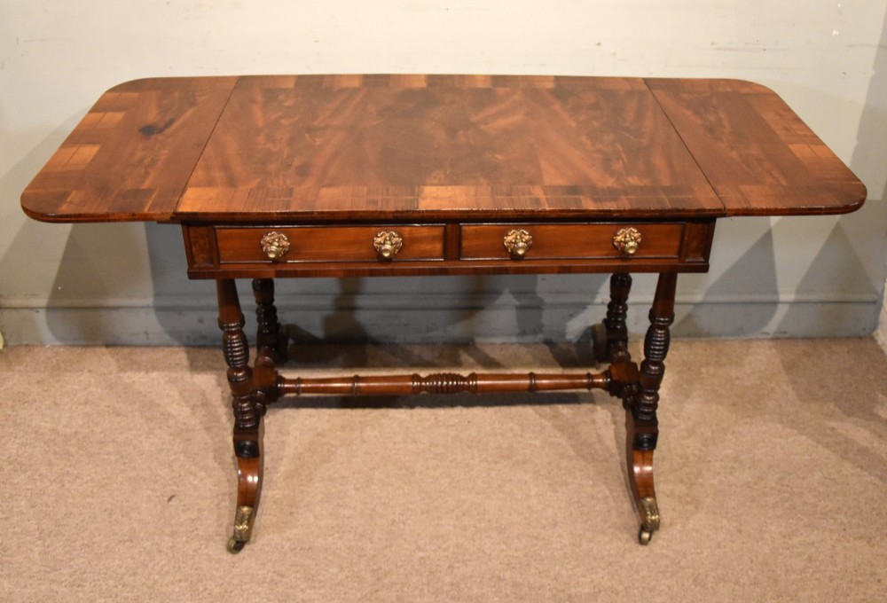 Regency flame mahogany sofa table featuring two drop leaves, two frieze drawers with brass handles, turned double supports, a central stretcher, and brass castors.