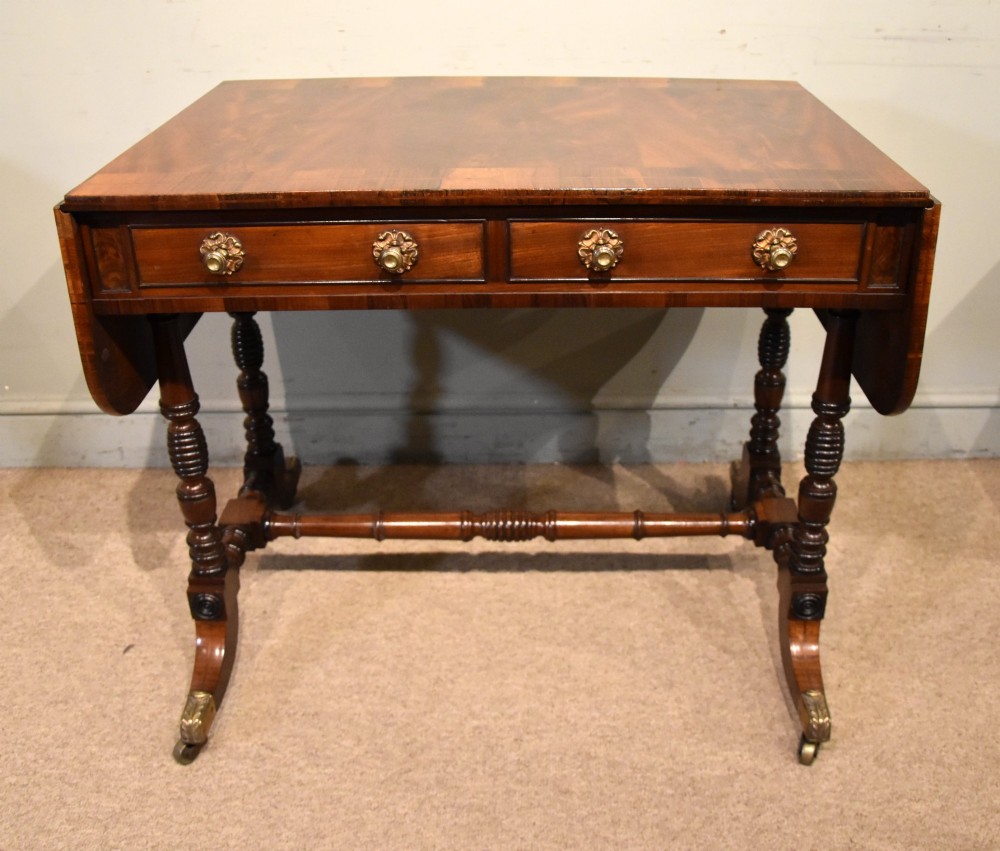 Regency flame mahogany sofa table featuring two drop leaves, two frieze drawers with brass handles, turned double supports, a central stretcher, and brass castors. View II