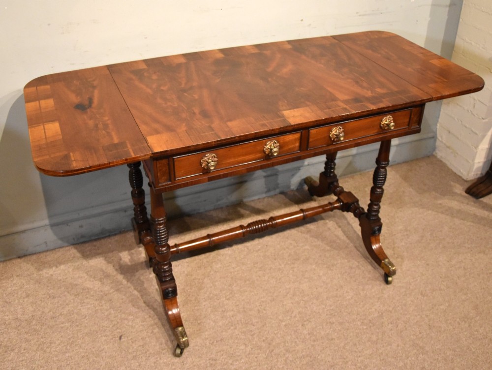 Regency flame mahogany sofa table featuring two drop leaves, two frieze drawers with brass handles, turned double supports, a central stretcher, and brass castors. View IV