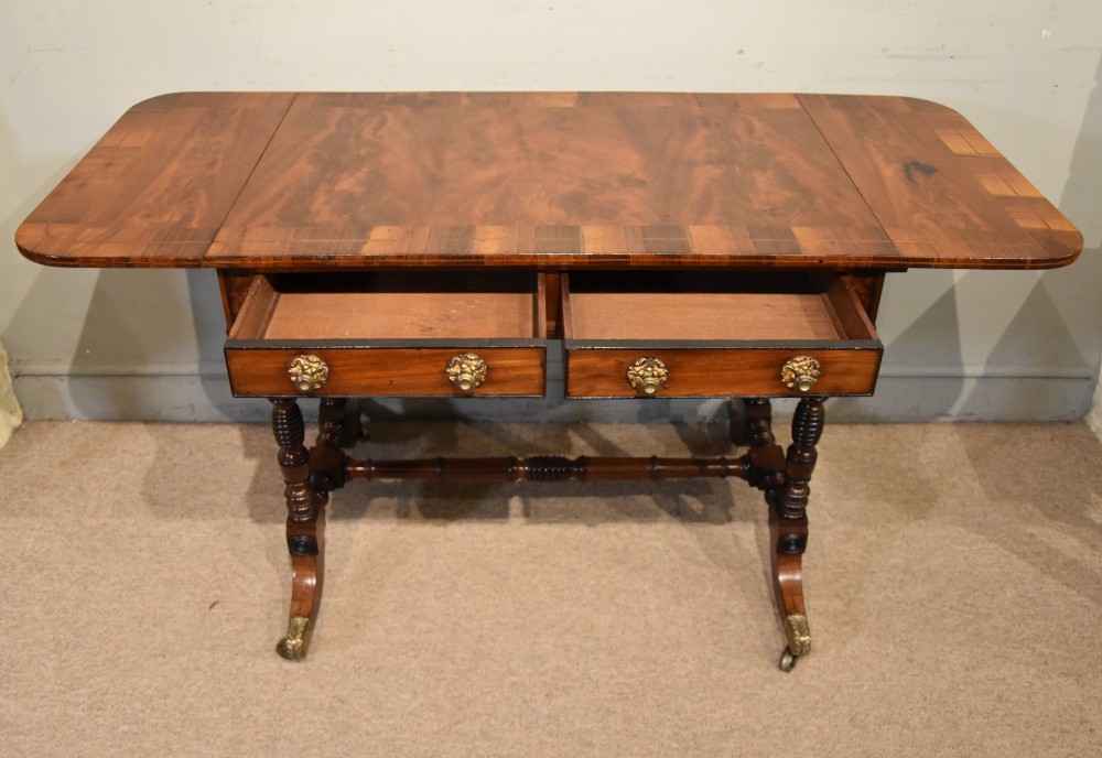 Regency flame mahogany sofa table featuring two drop leaves, two frieze drawers with brass handles, turned double supports, a central stretcher, and brass castors. View V
