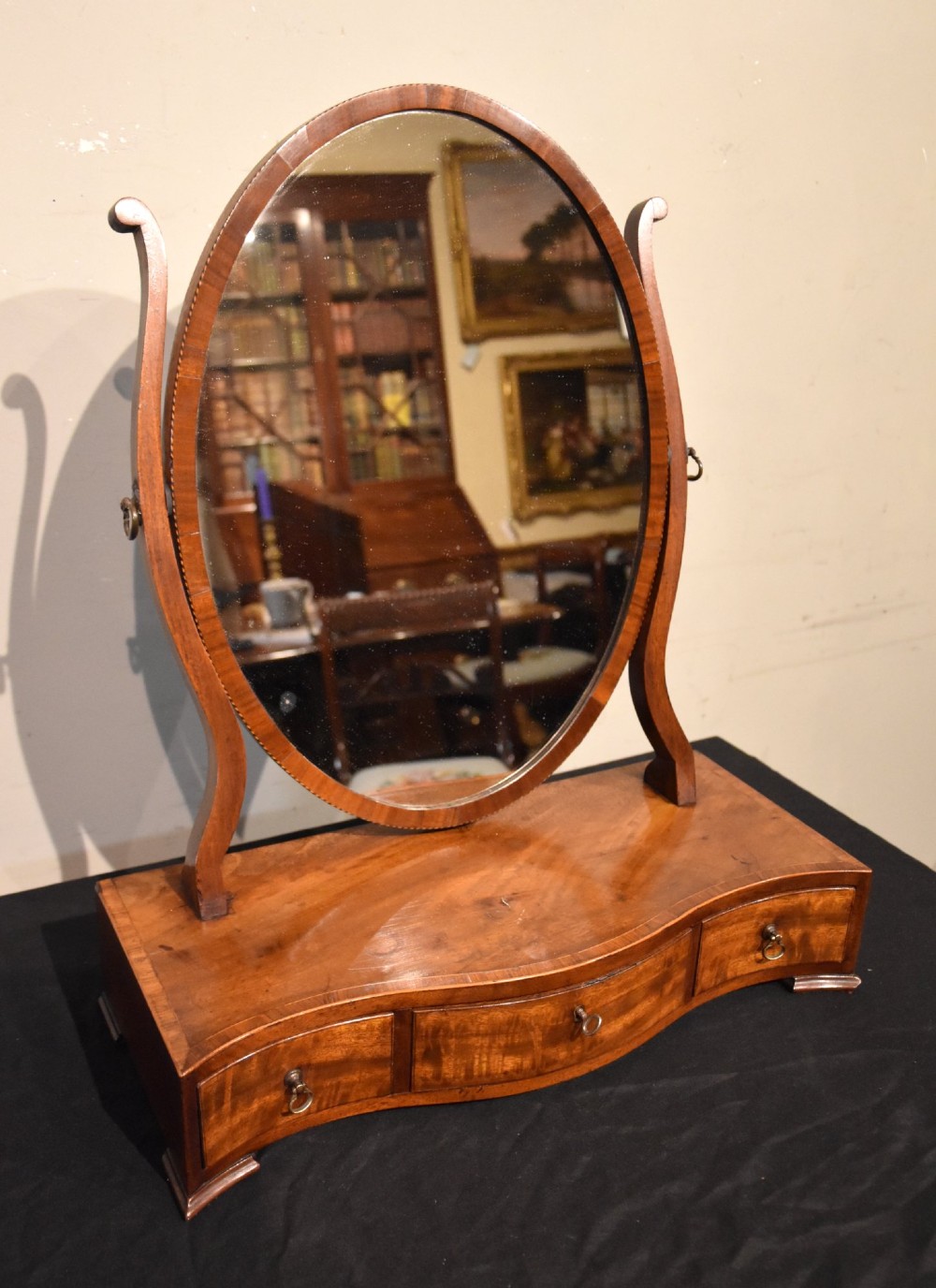 A Georgian mahogany dressing mirror featuring an oval adjustable mirror supported by curved uprights above a serpentine-fronted base with three small drawers.