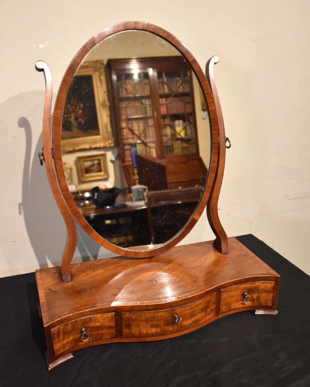 A Georgian mahogany dressing mirror featuring an oval adjustable mirror supported by curved uprights above a serpentine-fronted base with three small drawers. View II