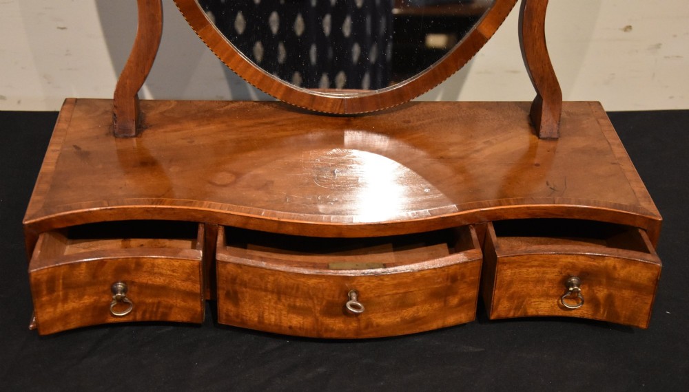 A Georgian mahogany dressing mirror featuring an oval adjustable mirror supported by curved uprights above a serpentine-fronted base with three small drawers. View IV