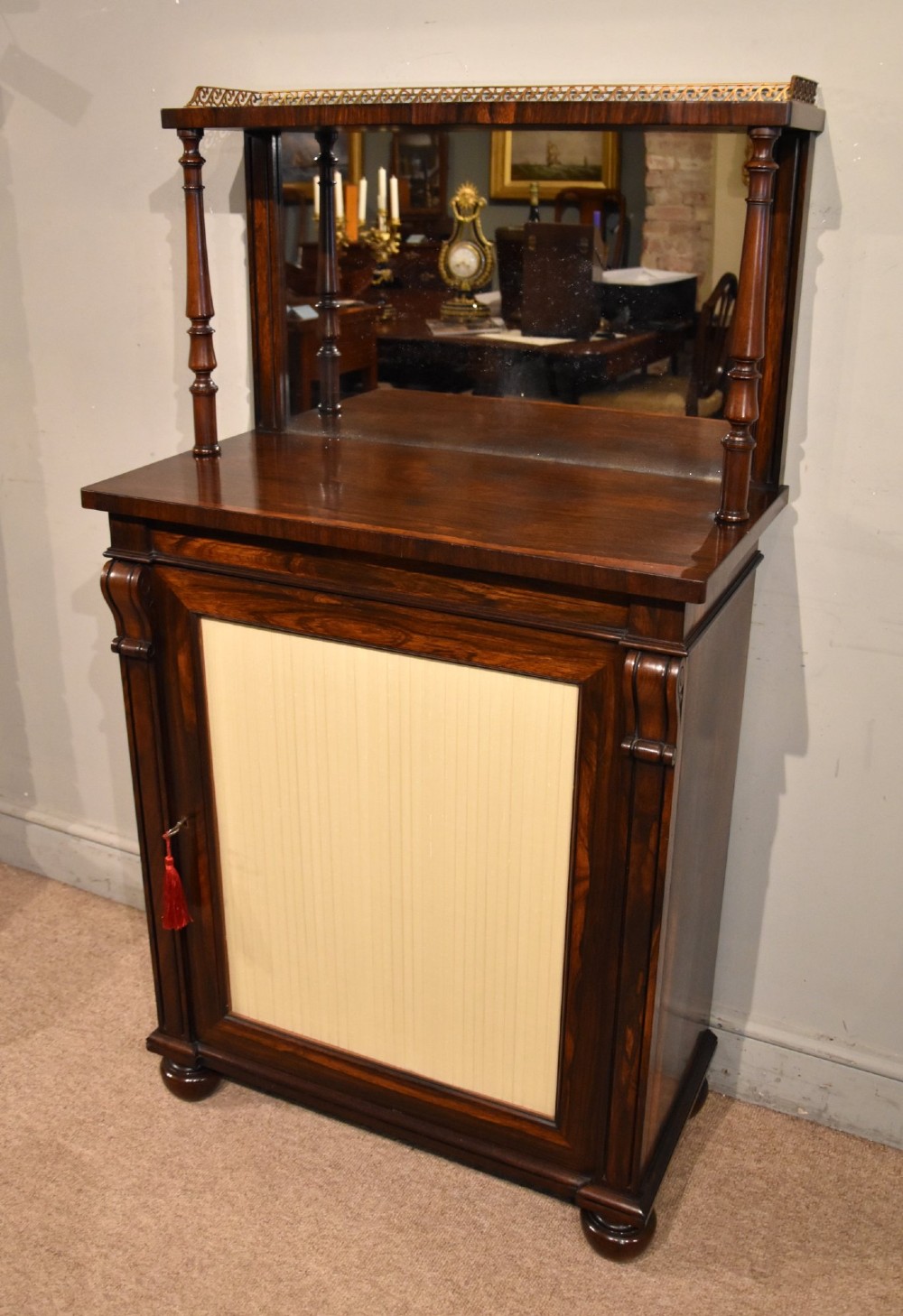 A George IV rosewood chiffonier featuring a raised mirrored back shelf with turned columns and a top brass gallery, set above a lower cupboard door with a pleated cream fabric panel. View II