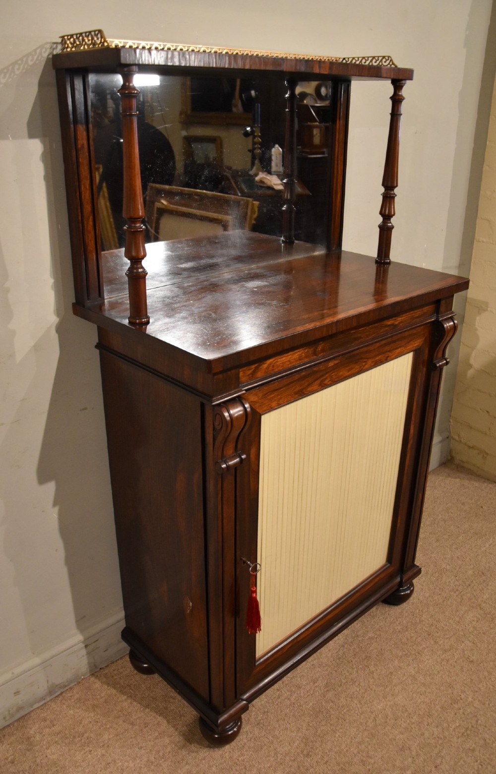 A George IV rosewood chiffonier featuring a raised mirrored back shelf with turned columns and a top brass gallery, set above a lower cupboard door with a pleated cream fabric panel. View VI