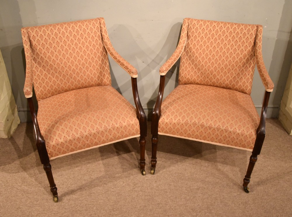 A pair of antique mahogany armchairs featuring terracotta pattern upholstery, sweeping armrests, turned front legs, and brass castors.